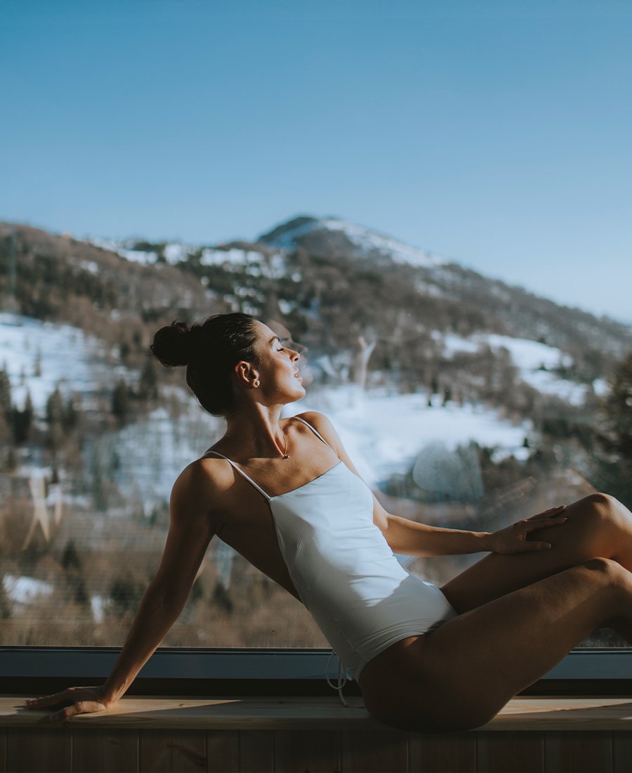 Donna in costume da bagno che si rilassa vicino a una finestra con vista sulle montagne innevate.
